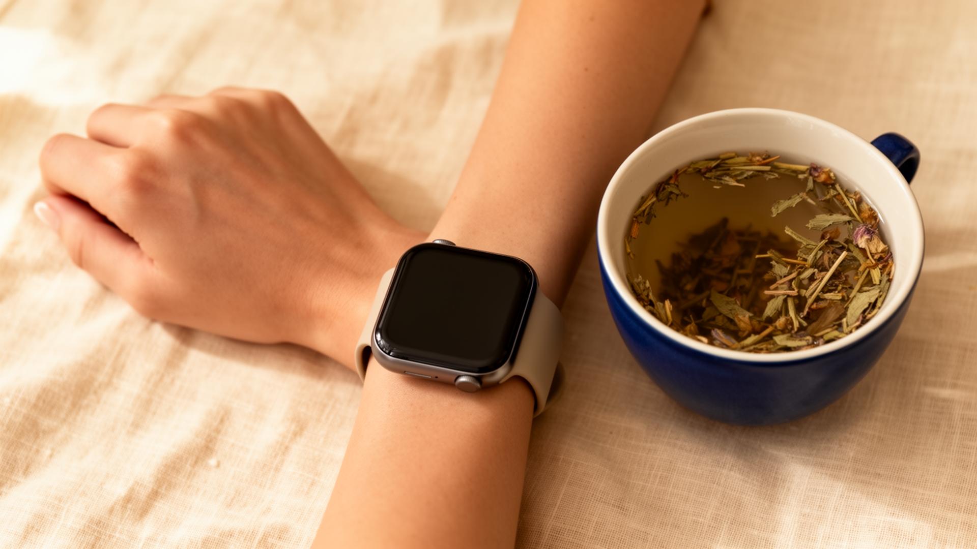 A woman's wrist wearing a smartwatch beside a cup of herbal tea on warm linen, a calm morning wellness scene